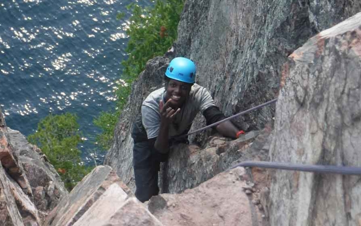 a person looks up and smiles at the camera while rock climbing above a body of water